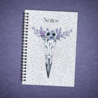 Carnet de notes à spirales noires représentant un crâne d'oiseau avec une couronne de fleurs et de perles violettes sur un fond gris clair avec des écritures calligraphiées